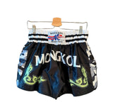 Black and blue camouflage Muay Thai shorts with green snake graphics and white 'MONGKOL' lettering, hanging on a wooden hanger against a dark smoky background. Text at the top reads 'NEW DROP' and at the bottom 'MONGKOL CAMO EDITION'.
